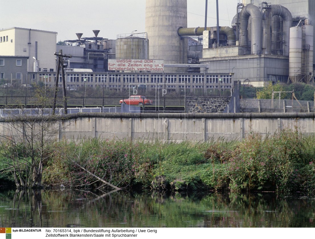 Innerdeutsche Grenze 1984-1987, Blick auf eine Mauer und einen Stahlgitterzaun mit einem großen Plakat "UdSSR und DDR - für alle Zeiten eng und brüderlich verbunden", dahinter das Zellstoffwerk<BR>Aufnahmedatum: 1984 - 1987<BR>Material/Technik: Positiv farbe<BR>4,2 cm x 5,7 cm<BR>Inventar-Nr.: 36/1<BR>