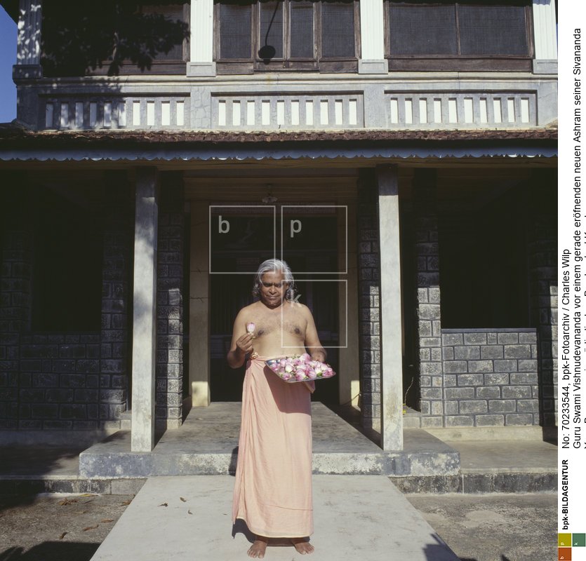 In indischer Tradition bietet er als Hausherr den eintretenden Gästen ein „Prasad“ an, kleine Geschenke und Blumen. Vishnudevananda organisierte eines der ersten Yoga-Lehrer-Trainingsprogramme in der westlichen Welt, den Sivananda Yoga-Lehrer-Kurs. Er gilt als Vertreter des Hatha und Raja Yoga.<BR>Aufnahmedatum: 1978<BR>Material/Technik: Foto<BR>Aufnahmeort: Indien<BR>