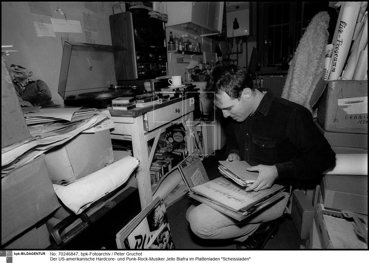 1980 eröffnete Norbert Hähnel seinen Plattenladen Scheissladen in Berlin-Kreuzberg. Er gehörte mit wenigen anderen Läden zu den Vertriebsorten einer Untergrund-Musik von Punk, experimenteller Musik bis zur Neuen Deutschen Welle.<BR>Person: Jello Biafra (bürgerlich Eric Reed Boucher (1958-), US-amerikanischer Hardcore- und Punk-Rock-Musiker sowie politischer Aktivist, der als Leadsänger der Punk-Band Dead Kennedys Bekanntheit erlangte.

Norbert Hähnel (1951-), deutscher Sänger aus Berlin-Kreuzberg. Er war der Inhaber des Plattenladens Scheissladen. Außerdem war er eine Zeit als Veranstalter von Punk-Konzerten tätig.<BR>Aufnahmedatum: 10.12.1982<BR>Material/Technik: Foto<BR>Aufnahmeort: Berlin-Kreuzberg (Berlin West)<BR>