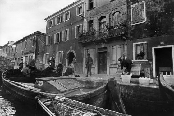 Szenerie am Kanal in Venedig. Eine Gruppe von Männern sitzt auf der Straße und dem angelegten Boot und flickt ein Fischernetz. Daneben wäscht eine Person Geschirr in großen Schüsseln. Sie wird von der Männergruppe und anderen Leuten auf der Straße beobachtet.