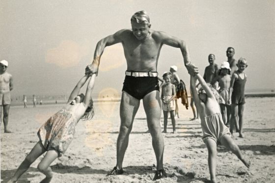 Am Strand - Ein kräftiger Mann hebt an jeder Hand ein Kind hoch
